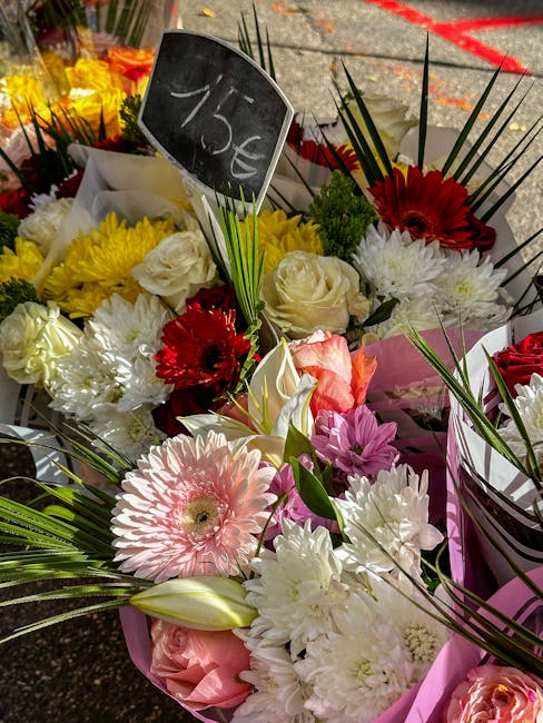 A vibrant floral arrangement featuring a variety of flowers including large white roses, pink gerbera daisies, red gerberas, and white chrysanthemums, all wrapped in pink paper and displayed outdoors 