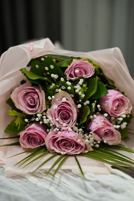 A close-up of a bouquet featuring nine soft pink roses with tightly layered petals, accented by small white baby's breath flowers and lush green foliage. The roses are arranged in a natural, rounded s