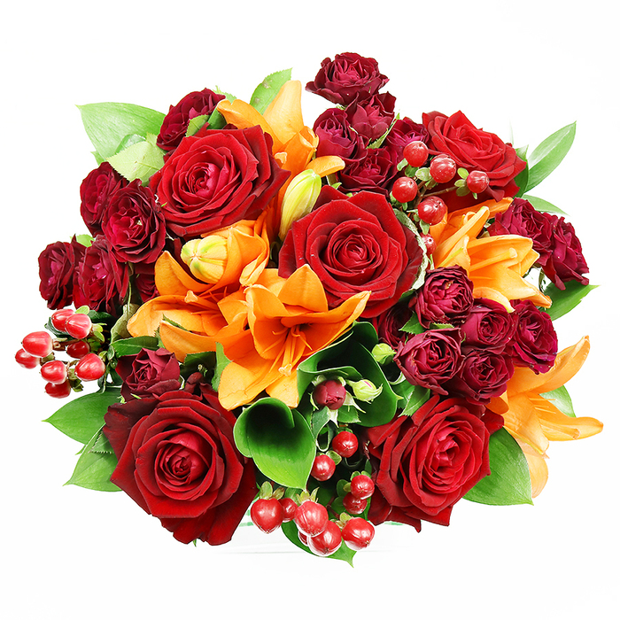 Captured straight overhead on a clean white background, this densely packed bouquet is a study in warm, jewel-like colour and careful floral craft. Several large, fully opened deep red roses anchor the design, their velvety petals layered to create a sense of timeless romance and to suggest a soft, familiar rose scent if drawn close. Interspersed are bright orange Asiatic lilies, some trumpet-shaped and fully blossomed with dark freckles and visible stamens, others held as tight buds that promise continuing bloom - a thoughtful touch that keeps the arrangement lively across days. Tight clusters of deep burgundy spray roses add concentrated colour and velvety texture, their miniature blooms creating shadowed pockets that deepen the palette. Glossy Hypericum berries in vivid ruby tones are dotted throughout, offering shiny, festive highlights and a contrast in shape and scale to the softer blossoms. Broad, glossy green leaves-likely ruscus or salal-peek and fan between the flowers, supplying a cool, verdant backdrop that lifts the warm reds and oranges. The lighting is even and gentle, accentuating the tactile differences from satiny lilies to plush roses and the smooth skins of the berries. As a local arrangement from Creekmouth Florist, this bouquet feels at home for occasions around Creekmouth and neighbouring Barking: an anniversary, a celebratory dinner, or a seasonal gift that reads as generous and warmly expressive. It balances drama with approachability, designed to sit beautifully on a dining table or to be presented as a confident, heartfelt gesture.